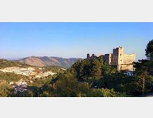 Panorama collinare con una grande fortezza in pietra in cima a un’altura, circondata da boschi e alberi. Sullo sfondo si vedono montagne e un piccolo centro abitato con case sparse lungo la valle sotto un cielo sereno.