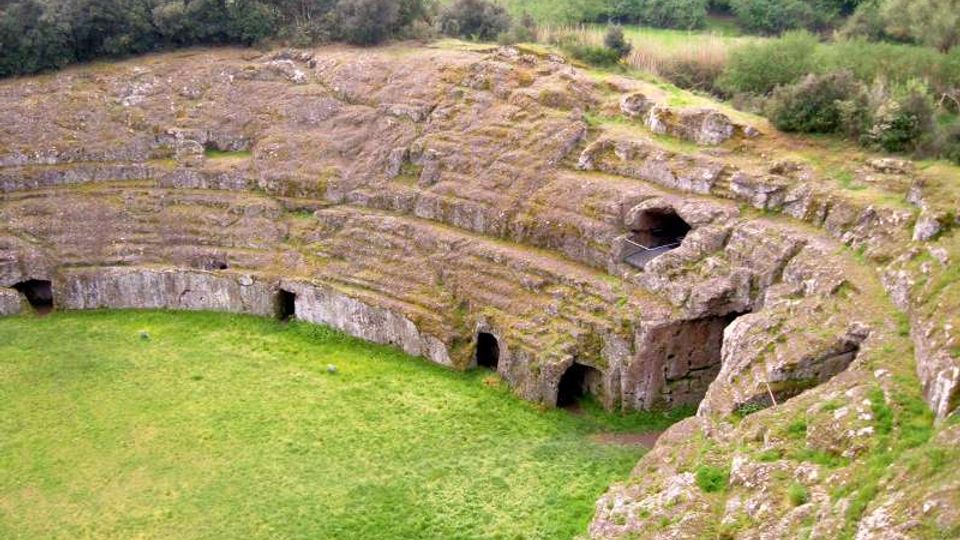 Anfiteatro antico scavato nella roccia, con gradinate semicircolari in pietra e piccole aperture ad arco lungo il perimetro. Al centro si vede un’area erbosa, circondata da vegetazione e colline sullo sfondo.