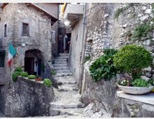 Vicolo stretto in un borgo in pietra con ripida scalinata di gradini irregolari tra muri e case in muratura, con lampione sospeso e cavi a vista. Piante in vaso e una piccola fioriera su muretto di pietra, con ingresso ad arco e bandiera italiana su una facciata.