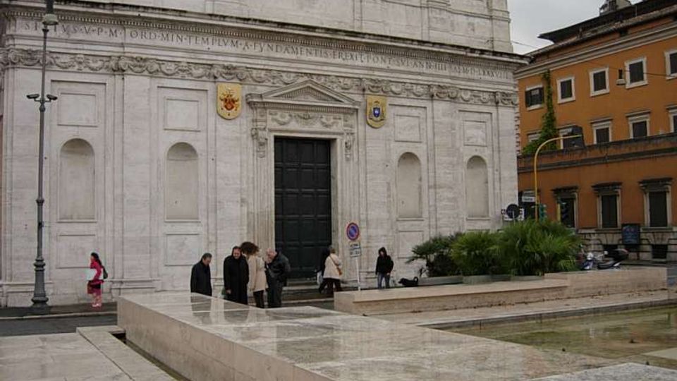 Facciata in pietra chiara di una chiesa monumentale con grande portale centrale, cornici scolpite e iscrizione lungo il fregio. In primo piano una piazza pavimentata con muretti bassi e una vasca d’acqua rettangolare; sullo sfondo edifici urbani e lampioni.