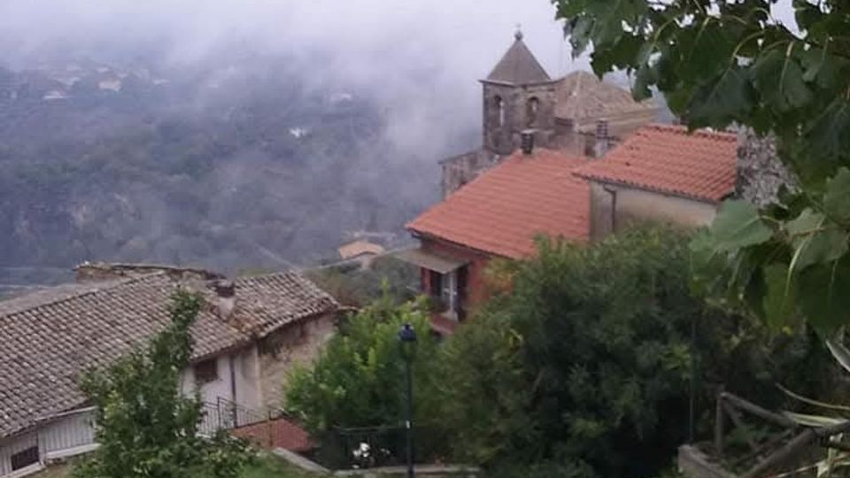 Vista di un borgo collinare con tetti in tegole rosse e una piccola chiesa in pietra con campanile, affacciati su una valle avvolta dalla nebbia. In primo piano si vedono un sentiero in ciottoli, staccionate in legno, vegetazione e rami di alberi che incorniciano la scena.