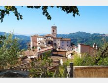 Veduta panoramica di un borgo collinare con case dai tetti in coppi e una torre campanaria con orologio al centro. In primo piano si vede una ringhiera e vegetazione, con montagne boscose sullo sfondo sotto un cielo sereno.