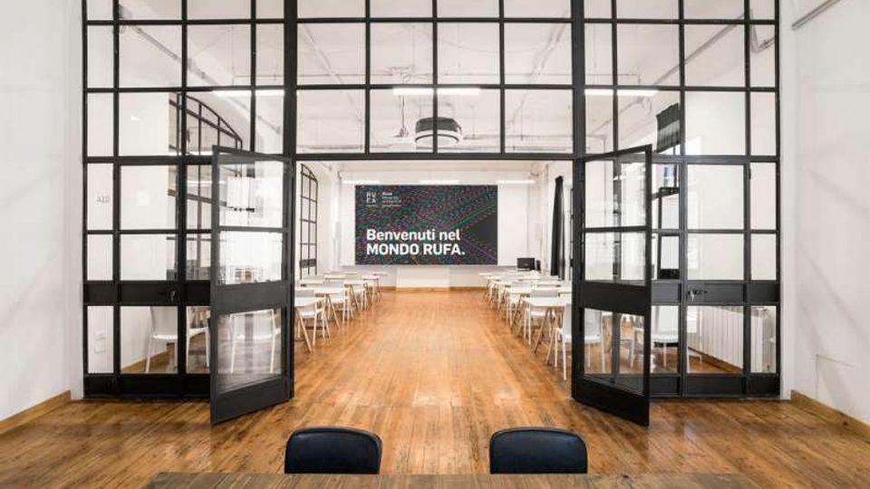 Ampia aula o spazio didattico con pavimento in legno, file di tavoli e sedie bianche ai lati e un grande cartello sulla parete di fondo. L’ingresso è delimitato da una grande parete vetrata a griglia con porte a battente nere, in un ambiente luminoso con soffitto alto e luci industriali.