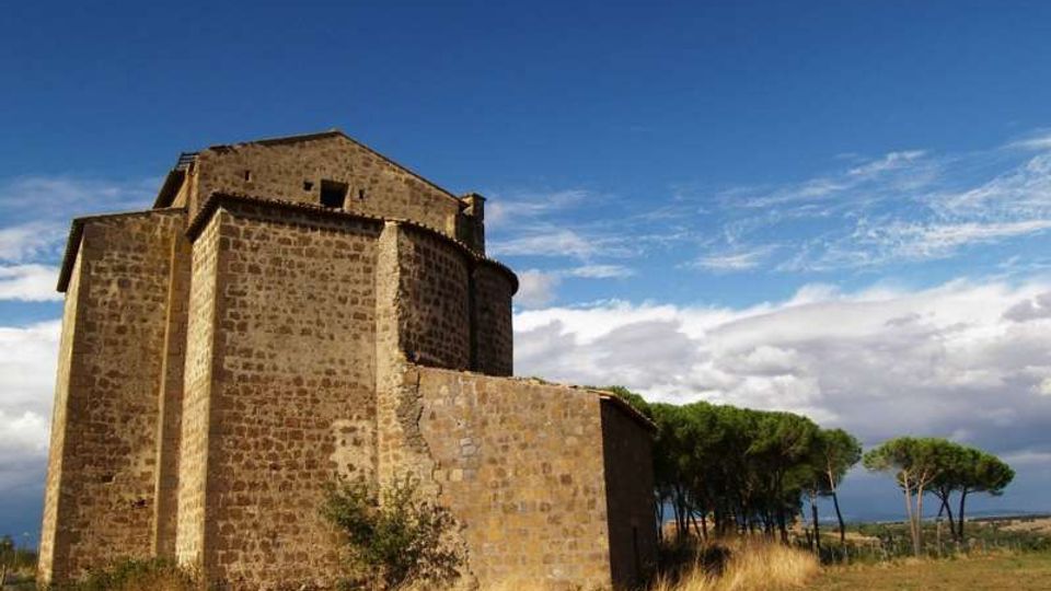 Chiesa in pietra di aspetto romanico con abside semicircolare e volume massiccio, situata in un’area rurale con terreno erboso secco. Sullo sfondo si vedono file di pini e un ampio cielo azzurro con nuvole.