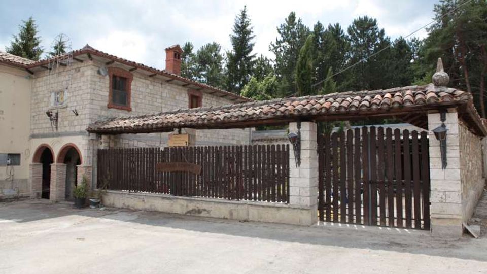 Edificio rurale in pietra a due piani con tetto in tegole e piccola loggia ad archi, affacciato su un cortile pavimentato. Davanti si vede una lunga recinzione e un grande cancello in legno con copertura in tegole, con alberi alti sullo sfondo.