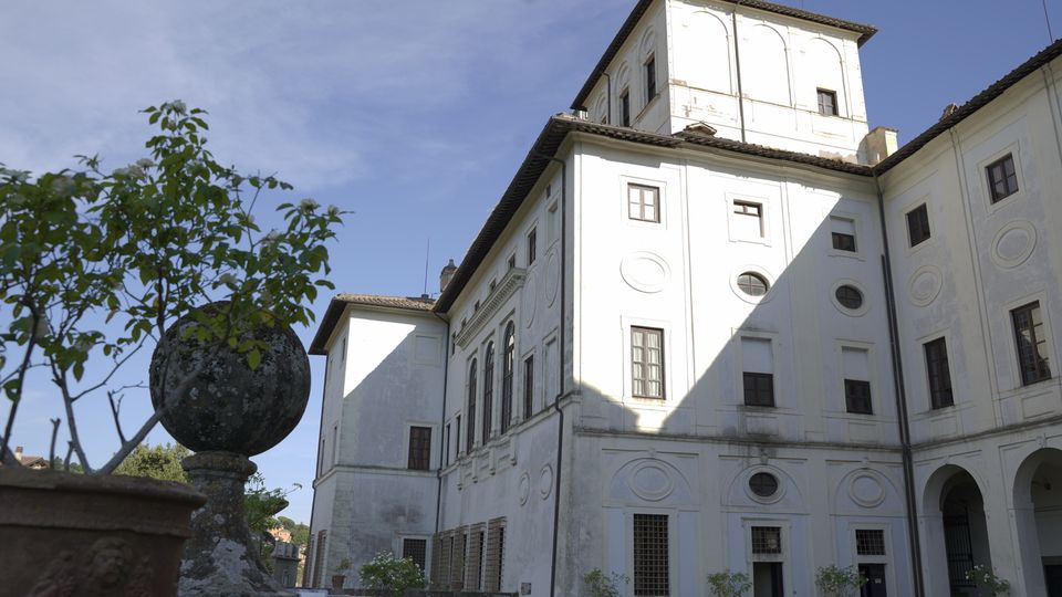 Facciata di un grande palazzo storico intonacato di bianco, con finestre ad arco e ovali decorativi, affacciato su un cortile con vasi e una balaustra in pietra. In primo piano si vede un vaso in terracotta e una sfera di pietra su un pilastro, con cielo sereno sullo sfondo.