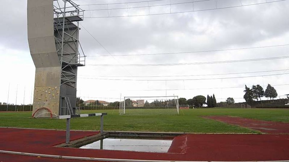 Campo sportivo all’aperto con pista di atletica rossa e prato centrale con porte da calcio. A sinistra si vede una torre alta per arrampicata o allenamento con struttura metallica, sotto un cielo nuvoloso e con cavi sospesi sopra l’area.