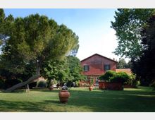 Ampio giardino con prato curato, grandi alberi e vasi in terracotta distribuiti sul terreno. Sullo sfondo si vede una casa rurale in mattoni rossi con finestre e pergolato, circondata dalla vegetazione.