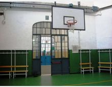Interno di una palestra scolastica con pavimento verde, pareti bianche e verdi e canestro da basket montato sopra una porta ad arco con grate metalliche. Lungo le pareti sono presenti panche e appendiabiti.