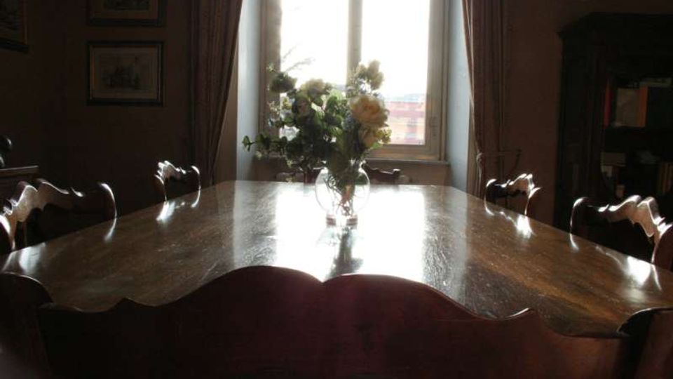 Sala da pranzo con grande tavolo ovale in legno lucido circondato da sedie intagliate, con un vaso di fiori al centro. Sullo sfondo una finestra luminosa con tende e alcune stampe incorniciate alle pareti, accanto a una libreria scura.