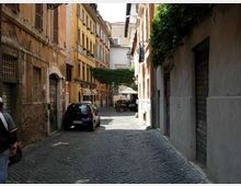 Strada stretta in acciottolato tra alti edifici storici con facciate color ocra e persiane, con un’auto parcheggiata sul lato sinistro. In fondo si vede una piccola piazzetta ombreggiata con alberi e tavolini sotto ombrelloni.