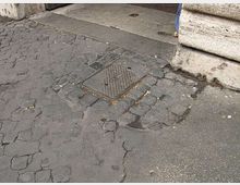 Marciapiede urbano con asfalto molto crepato e un tombino quadrato in metallo al centro. Sul lato destro si vede la base in pietra di un edificio e una soglia d’ingresso in pavimentazione liscia.