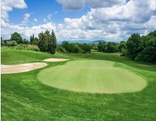 Campo da golf con ampio green in primo piano e bandierina al centro, circondato da fairway ondulati e bunker di sabbia sulla sinistra. Sullo sfondo si vedono alberi, colline e un cielo con nuvole sparse.