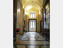 Interno di un corridoio in stile storico con soffitto a volta decorato e illuminazione a parete, pavimento in pietra con motivi geometrici e una finestra sul fondo. Sulla destra si vede una parete con grandi vetrate e porte, mentre a sinistra sono presenti colonne e un pannello informativo.