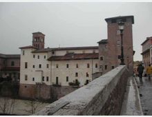 Vista da un ponte in pietra con parapetto e lampione su un complesso di edifici storici lungo un fiume, con facciate intonacate chiare e una torre in mattoni. Sullo sfondo si vede un campanile e i tetti in coppi sotto un cielo grigio.