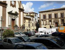 Piazza cittadina con parcheggio, circondata da edifici storici in pietra con persiane e facciate a più piani. A sinistra si vede una chiesa con portale monumentale e orologio sulla facciata, con lampioni e aiuole lungo il bordo della piazza.