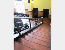 Scala interna in un edificio con gradini in rosso e corrimano in metallo, che scende verso un piano inferiore. Sullo sfondo si vedono una grande finestra a riquadri, pareti con rivestimento in marmo e due porte scure.