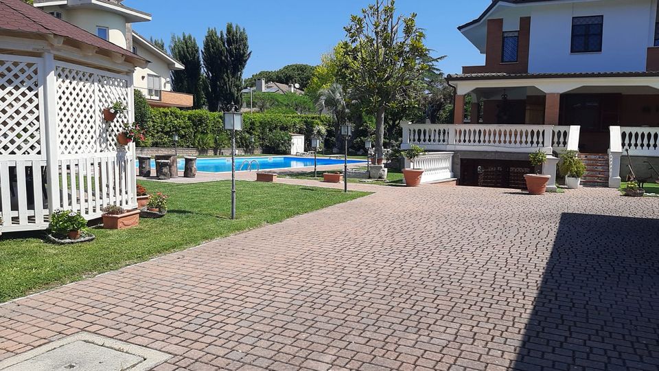 Cortile residenziale con ampia pavimentazione in autobloccanti e prato curato, con piscina rettangolare sullo sfondo. Ai lati si vedono una villetta a due piani con portico e balaustra bianca e un gazebo bianco in legno, circondati da siepi e alberi.