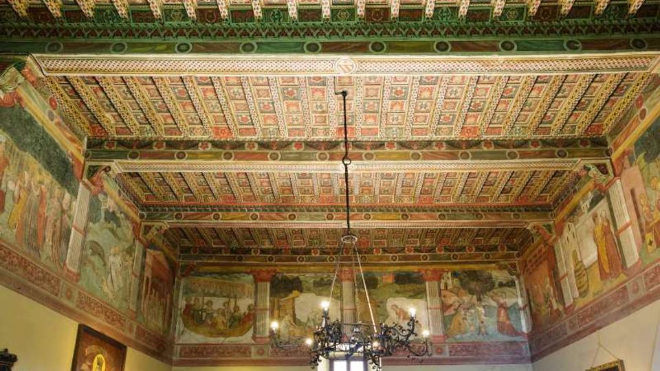 Interno di una sala storica con soffitto a cassettoni dipinto e travi decorate, e pareti affrescate con scene figurative lungo il perimetro superiore. Al centro pende un grande lampadario in ferro battuto sopra una finestra e quadri appesi alle pareti.