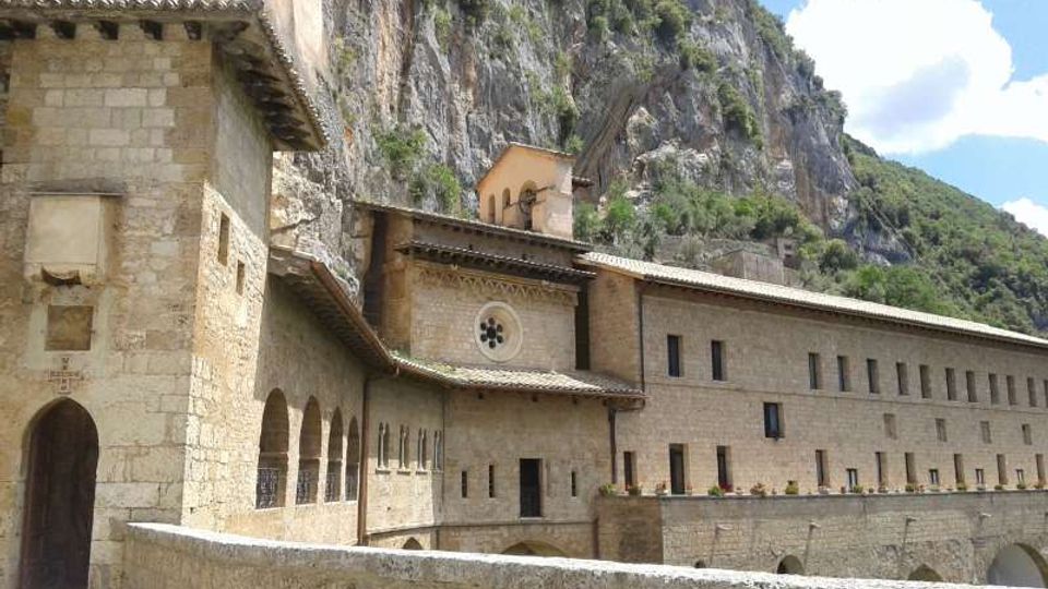 Complesso monastico in pietra con arcate, finestre regolari e piccolo campanile, addossato a una parete rocciosa. In primo piano un muretto e sullo sfondo una montagna boscosa sotto cielo parzialmente nuvoloso.