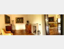 Interno di una casa con pavimento in cotto, pareti chiare e quadri incorniciati; al centro si vede un camino in pietra e mobili in legno. Una porta aperta conduce a una stanza adiacente arredata con divano e tavolo.