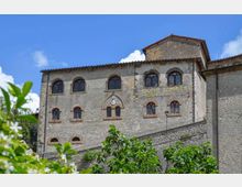 Facciata in pietra di un edificio storico a più piani, con numerose finestre ad arco e cornici in laterizio, sormontato da un tetto in tegole. In primo piano si vedono arbusti verdi e sullo sfondo un cielo azzurro con alcune nuvole.