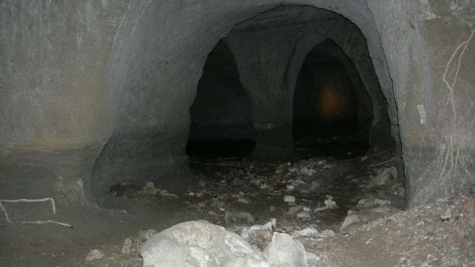Interno di una grotta o galleria sotterranea con pareti di roccia grigia e volte arrotondate sostenute da colonne naturali. Il pavimento è irregolare, coperto di terra e detriti con alcune pietre e massi in primo piano, e sullo sfondo si intravedono aperture più scure.