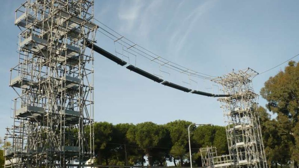 Due alte torri metalliche a traliccio con piattaforme e scale interne sostengono una passerella sospesa collegata da cavi. La struttura si trova all’aperto, con alberi sullo sfondo e edifici bassi nelle vicinanze sotto un cielo sereno.