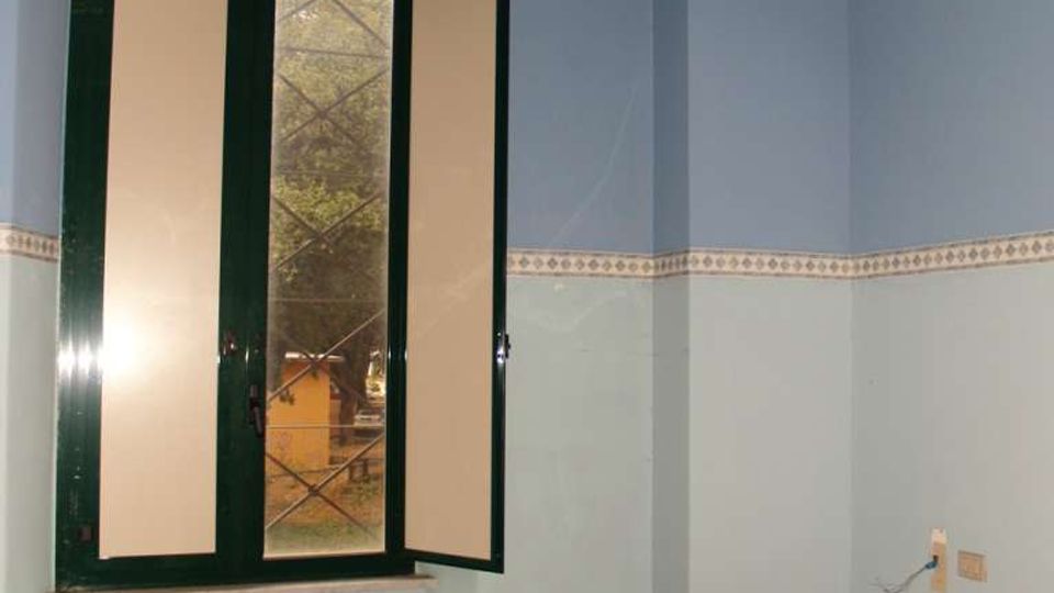 Interno di una piccola stanza vuota con pareti bicolore azzurro e fascia decorativa, pavimento piastrellato e zoccolino arancione. Finestra a due ante con telaio verde e una parte aperta, affacciata sull’esterno.