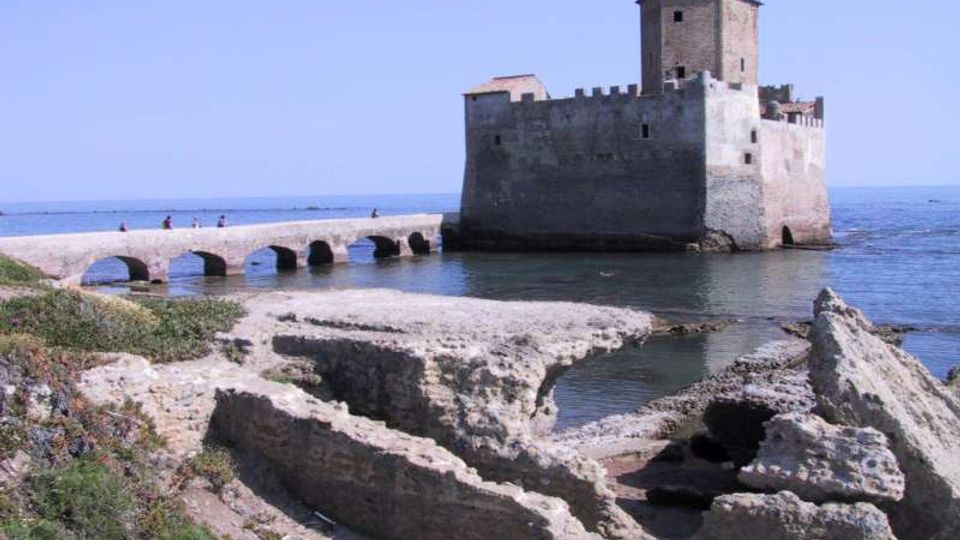 Fortezza in pietra sul mare con torre quadrata e mura merlate, collegata alla riva da un ponte in pietra ad archi. In primo piano si vedono rovine di murature e scogli lungo una piccola insenatura con acqua calma.