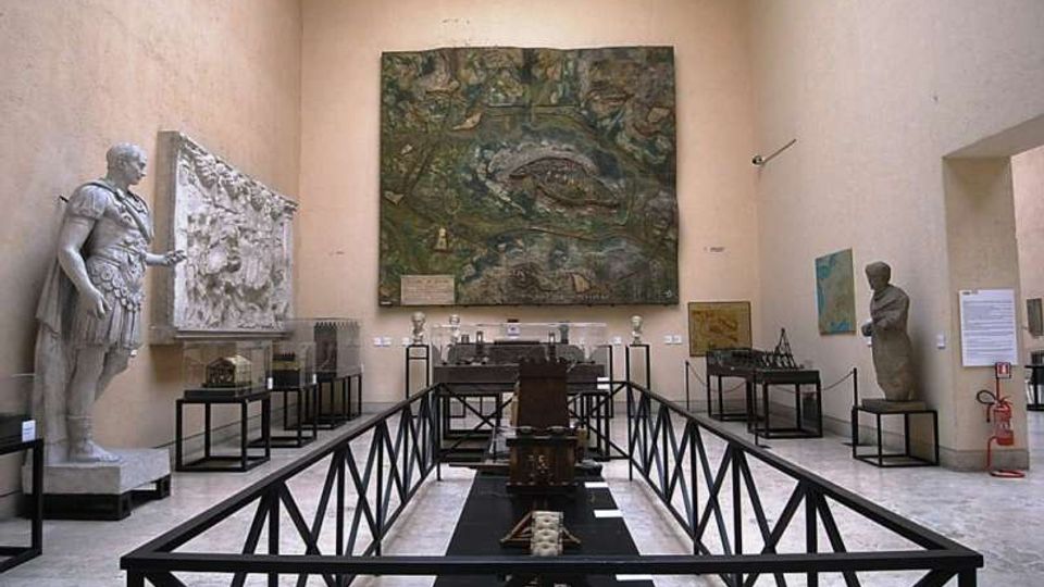 Sala museale con pareti alte color crema, opere d’arte e pannelli espositivi lungo i lati, tra cui grandi rilievi e statue. Al centro si vede un percorso delimitato da ringhiere nere con teche e modellini, e sulla parete di fondo un grande dipinto.