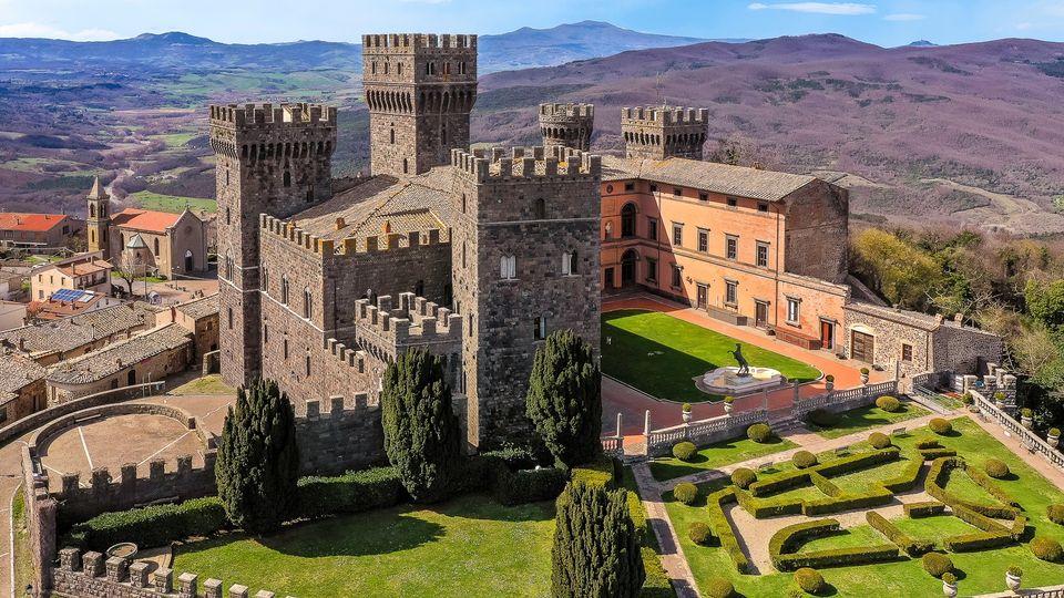 Veduta aerea di un castello medievale in pietra con torri merlate e mura fortificate, affiancato da un edificio in intonaco color ocra. Nel cortile interno e nei giardini geometrici con siepi potate si vede una fontana, con un borgo e colline montuose sullo sfondo.