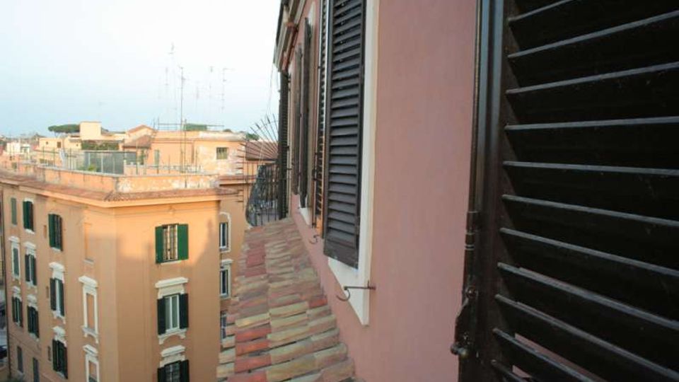 Vista da una finestra su un tetto inclinato in tegole e sulla facciata di un edificio color rosa con persiane nere. Sullo sfondo si vedono palazzi urbani color ocra con finestre e terrazze, sotto un cielo chiaro.