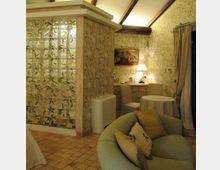 Interno di una camera o suite con arredi classici: pareti con carta da parati a motivi vegetali, pavimento in piastrelle e soffitto con travi a vista. Un divisorio in mattoni di vetro separa l’area soggiorno con poltrona imbottita, tavolino rotondo, credenza con lampada e grandi tende davanti a una porta-finestra.