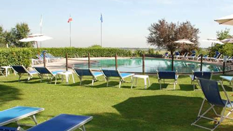 Piscina all’aperto circondata da prato con numerosi lettini blu e ombrelloni bianchi. Intorno si vedono siepi, alcuni alberi e pennoni con bandiere sullo sfondo sotto un cielo sereno.