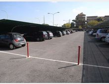 Parcheggio esterno asfaltato con file di auto sotto pensiline metalliche su entrambi i lati e corsia centrale ampia. Sullo sfondo si vedono lampioni stradali e alcuni edifici tra alberi.