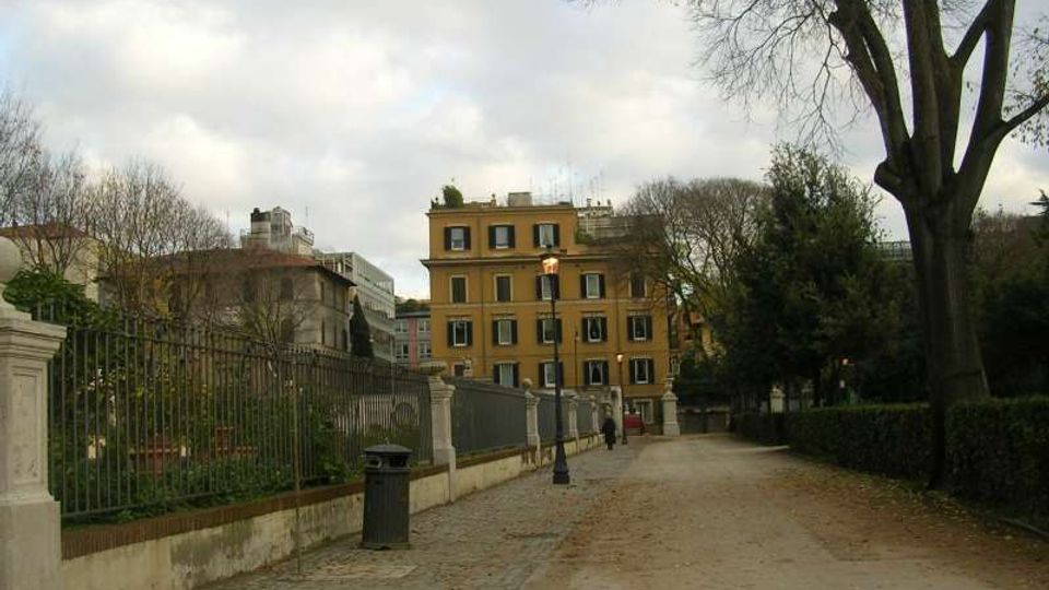 Viale pedonale in un parco urbano con sentiero in ghiaia e pavé, fiancheggiato da siepi, alberi e una recinzione in ferro battuto su muretto. Sullo sfondo si vede un edificio a più piani color ocra e lampioni lungo il percorso.