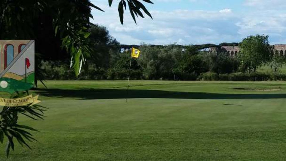 Campo da golf con green ben curato e bandierina gialla, circondato da prati e alberi. Sullo sfondo si vede un lungo acquedotto in pietra che attraversa il paesaggio sotto un cielo parzialmente nuvoloso.
