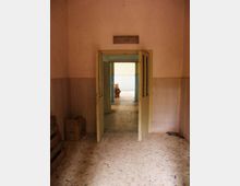 Interno di un edificio spoglio con pavimento in graniglia e pareti intonacate, con una doppia porta a battente aperta al centro. Attraverso l’ingresso si vede un corridoio con rivestimento a parete e luce naturale proveniente da un’apertura sul fondo.