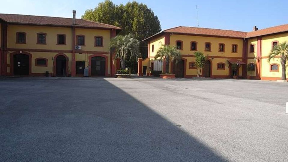 Cortile pavimentato e ampio circondato da edifici a due piani in stile rurale, con facciate gialle e bordi rossi, tetti in tegole e arcate al piano terra. Sullo sfondo si vedono alberi alti e alcune palme vicino agli ingressi.