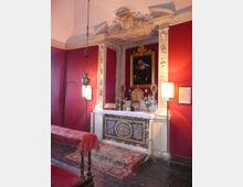 Interno di una piccola cappella con pareti rosso scuro e altare in marmo decorato, incorniciato da stucchi bianchi e sormontato da un dipinto religioso. Sul banco dell’altare sono presenti candelabri, fiori e libri, con lampade a parete e una lampada sospesa che illuminano la scena.