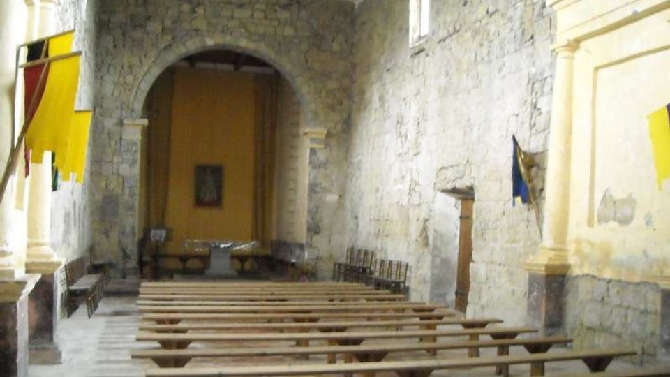 Interno di una piccola chiesa in pietra con navata semplice, file di panche in legno e grande arco che conduce all’area dell’altare sul fondo. Pareti in muratura a vista, finestre alte e soffitto con travi lignee.