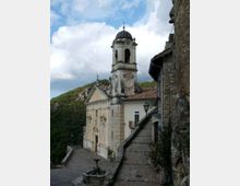 Chiesa storica in pietra e intonaco con facciata decorata e campanile con cupola scura, affacciata su una piccola piazza con fontana. Accanto corre una stretta strada lastricata lungo un muro in pietra, con colline boscose sullo sfondo sotto un cielo nuvoloso.