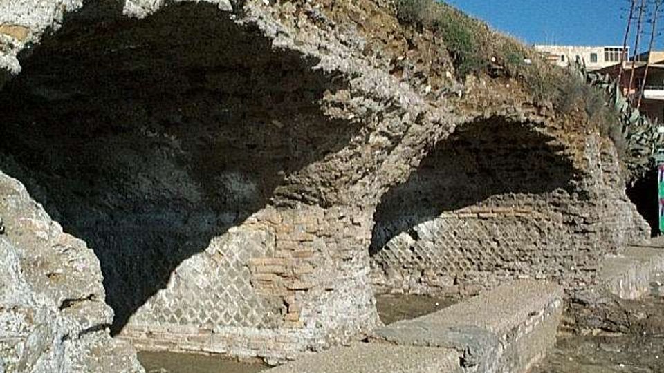 Resti di strutture in muratura con grandi archi in mattoni incassati in un pendio roccioso, affacciati su un’area pavimentata con vasche rettangolari. Sullo sfondo si vede cielo sereno e alcune costruzioni moderne in cima alla scarpata.