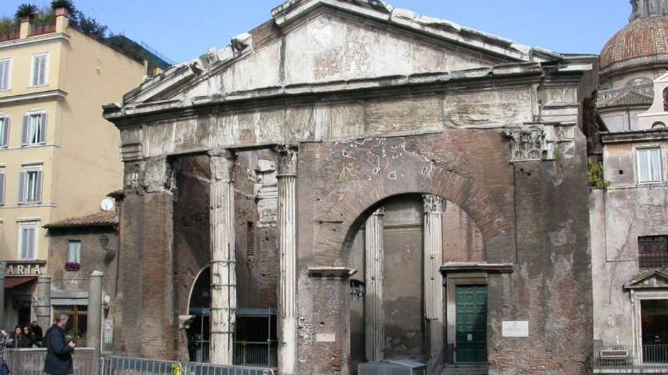 Facciata di un edificio storico in muratura e pietra, con frontone triangolare, grandi archi murati e colonne antiche parzialmente conservate. Davanti si vede una piazza con transenne metalliche e, sullo sfondo, palazzi adiacenti e una cupola visibile a destra.