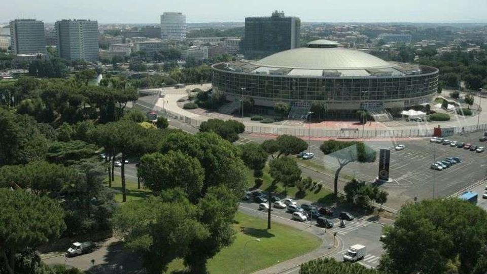 Vista aerea di un grande edificio moderno a pianta circolare con cupola bassa e facciata vetrata, circondato da strade e parcheggi. Intorno si estendono aree verdi con alberi e, sullo sfondo, palazzi e grattacieli della città.