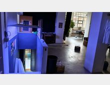 Interno di un loft o studio industriale con pavimento in cemento e pareti bianche, con un mezzanino e una scala che scende a un livello inferiore. Sullo sfondo grandi finestre a griglia illuminano una zona con tavolo e attrezzature, con colonne, tende nere e oggetti sparsi nello spazio.