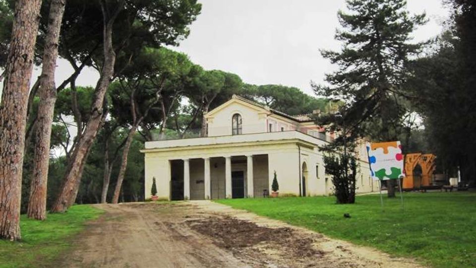 Edificio basso color crema con portico a colonne, situato in un’area verde alberata con alti pini. Un ampio viale sterrato conduce all’ingresso, con cartelloni informativi sul prato a destra.