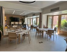 Sala riunioni moderna con tavoli rotondi e sedie beige su pavimento chiaro, illuminata da luci lineari a soffitto e con una parete in legno con schermo. Grandi porte-finestre in legno si aprono su un terrazzo con piante e vista su pareti in mattoni.