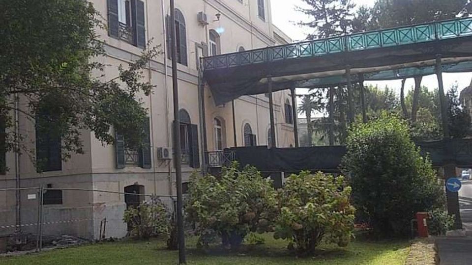 Edificio chiaro a più piani con finestre ad arco e persiane scure, affacciato su un prato con cespugli e alberi. A lato passa una passerella sopraelevata con parapetto verde, vicino a un vialetto asfaltato e a un lampione.
